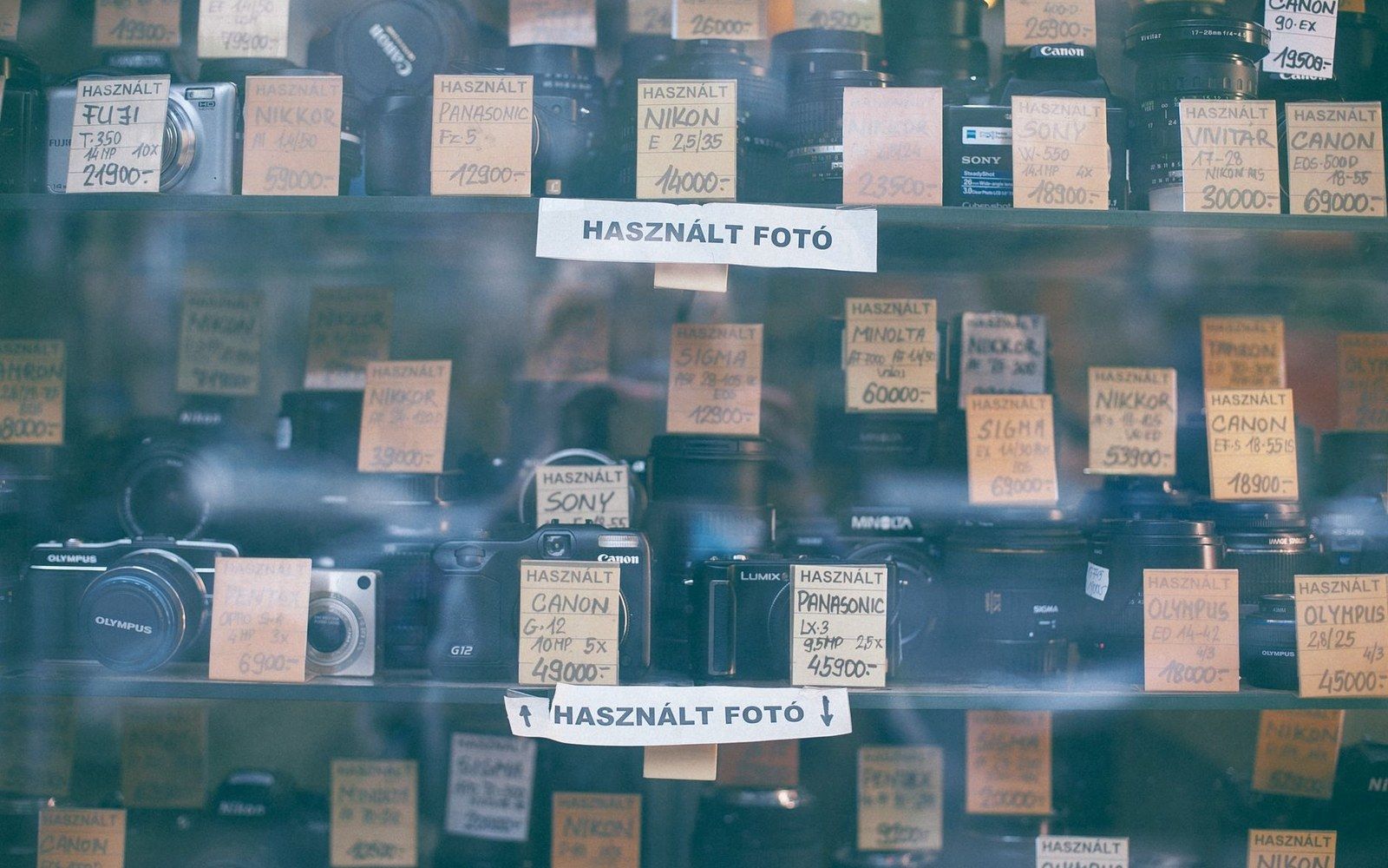 The window of a camera shop with used bodies and lenses displayed on shelves, street reflection visible in the glass, a person browsing inside