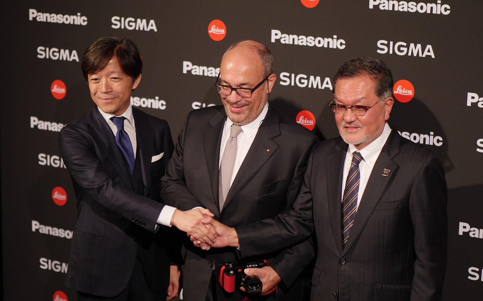 Three executives shake hands on stage in front of a Panasonic, Sigma and Leica backdrop