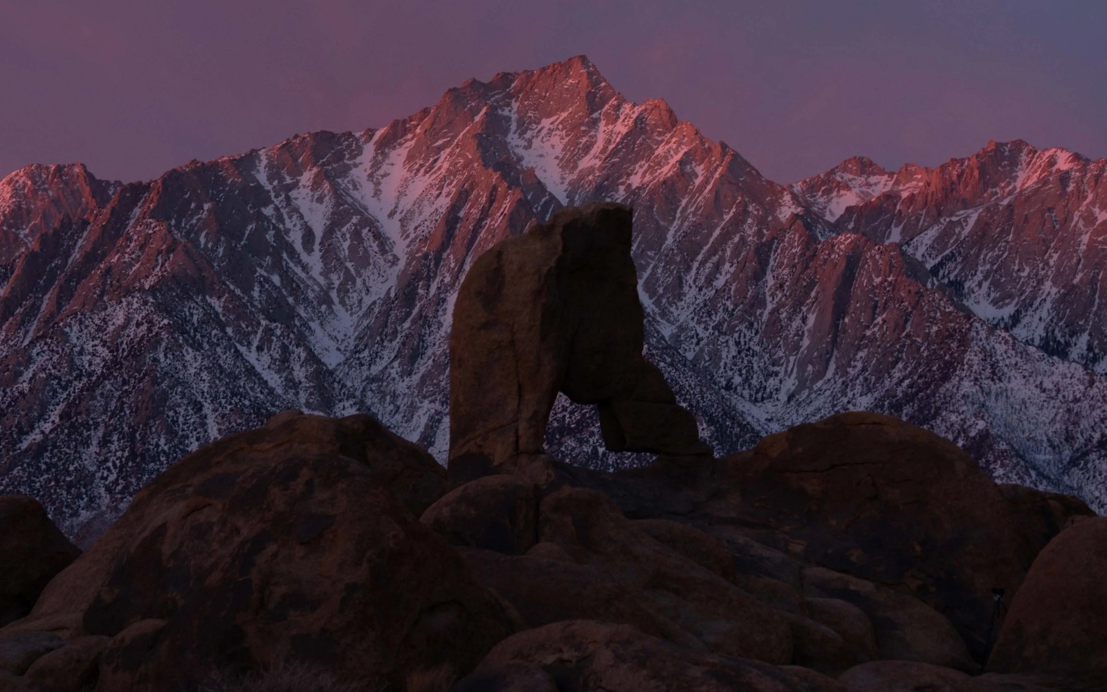 Silhouetted rock arch stands before snow-streaked mountain peaks glowing pink at dawn