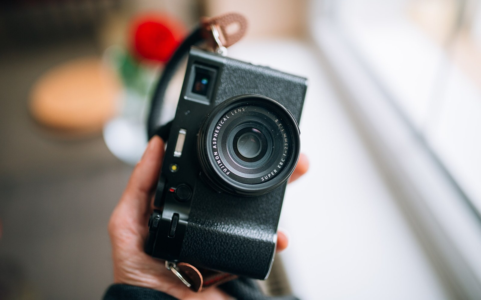 A hand holds a Fujifilm X100V camera, showing its fixed lens and leather strap